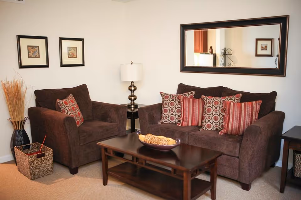 A cozy living room area with a dark brown armchair and sofa adorned with patterned red and beige throw pillows. A wooden coffee table with a decorative bowl sits in front of the sofa. On the wall above the sofa is a large rectangular mirror, and two framed pictures hang on the adjacent wall. A table lamp with a white shade is placed on a small side table between the armchair and sofa. A woven basket and a tall vase with dried decorative grass are positioned on the floor next to the armchair.