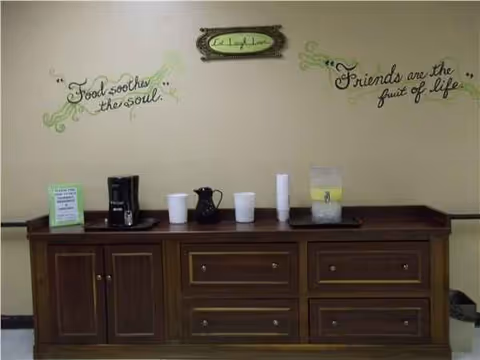 A wooden beverage counter with coffee maker, cups, and a pitcher against a wall with quotes.