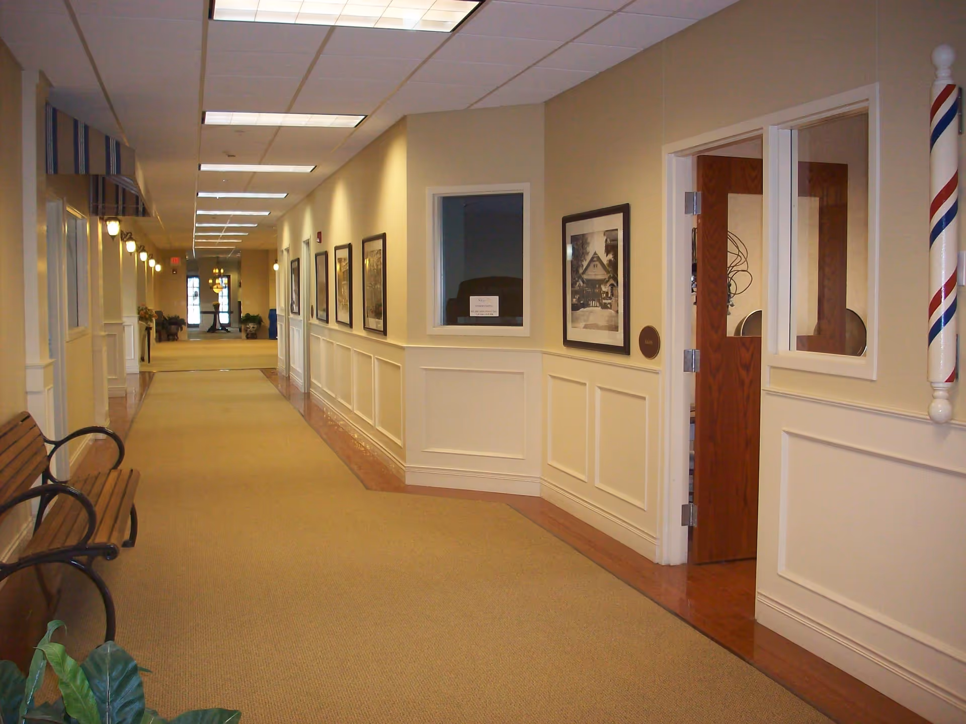 A long, well-lit interior hallway in a senior living facility with benches, framed pictures, doors, and a barber pole visible.