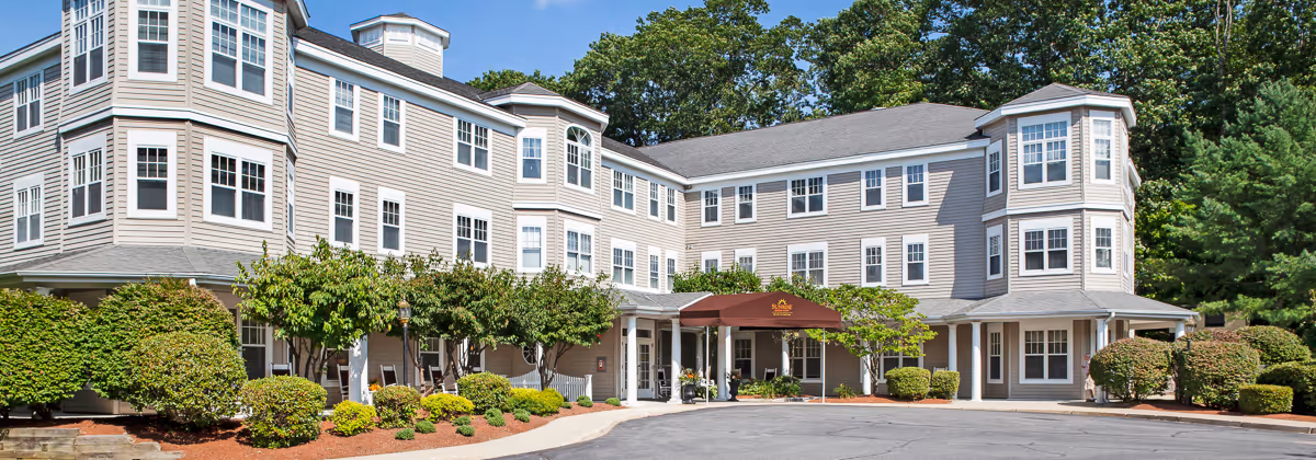 Exterior view of a three-story senior living facility building with beige siding, multiple windows, and a covered entrance with a maroon awning. The building is surrounded by well-maintained landscaping including bushes and small trees, with a paved driveway in front.