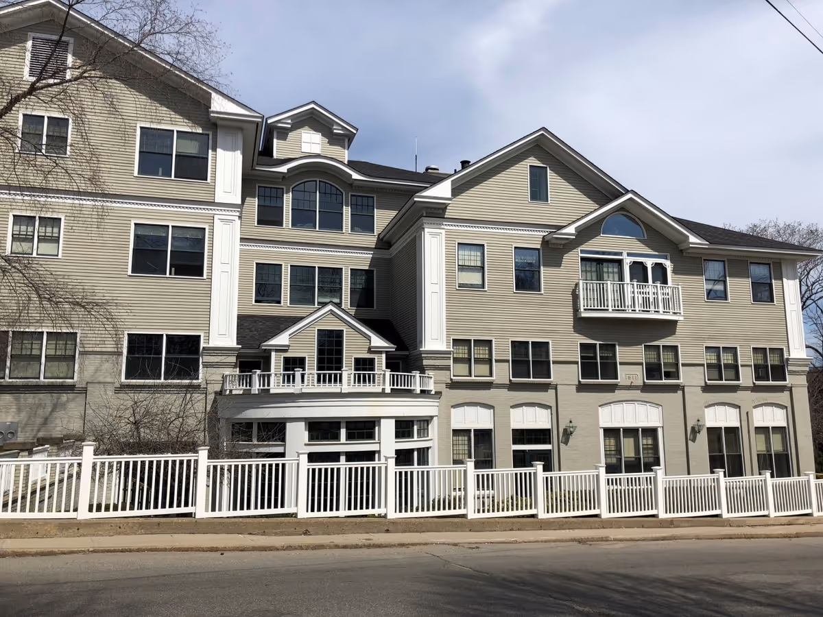 Exterior view of a multi-story senior living facility building with beige siding, multiple windows, a small balcony, and a white fence along the sidewalk in front.
