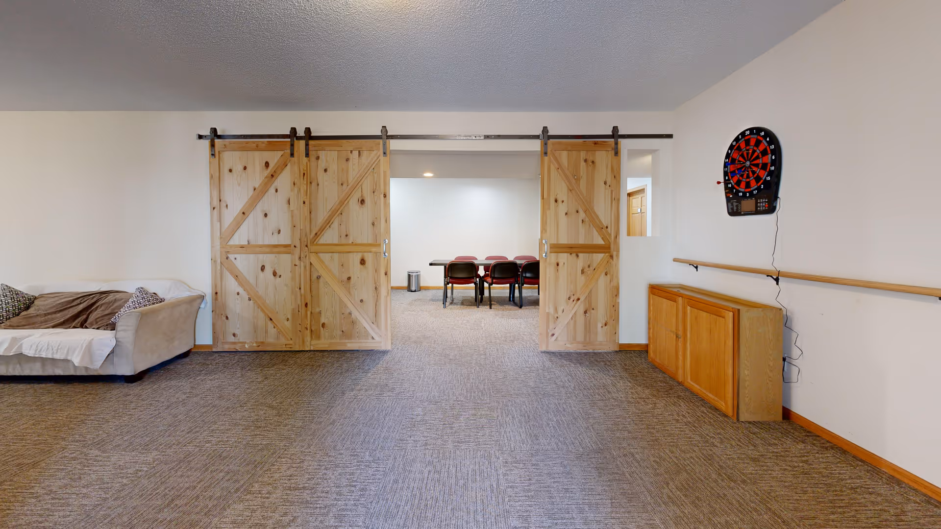 A spacious room with carpeted floor featuring a beige couch with pillows on the left, wooden sliding barn doors in the center leading to a room with a table and chairs, and a dartboard mounted on the right wall above a wooden cabinet.