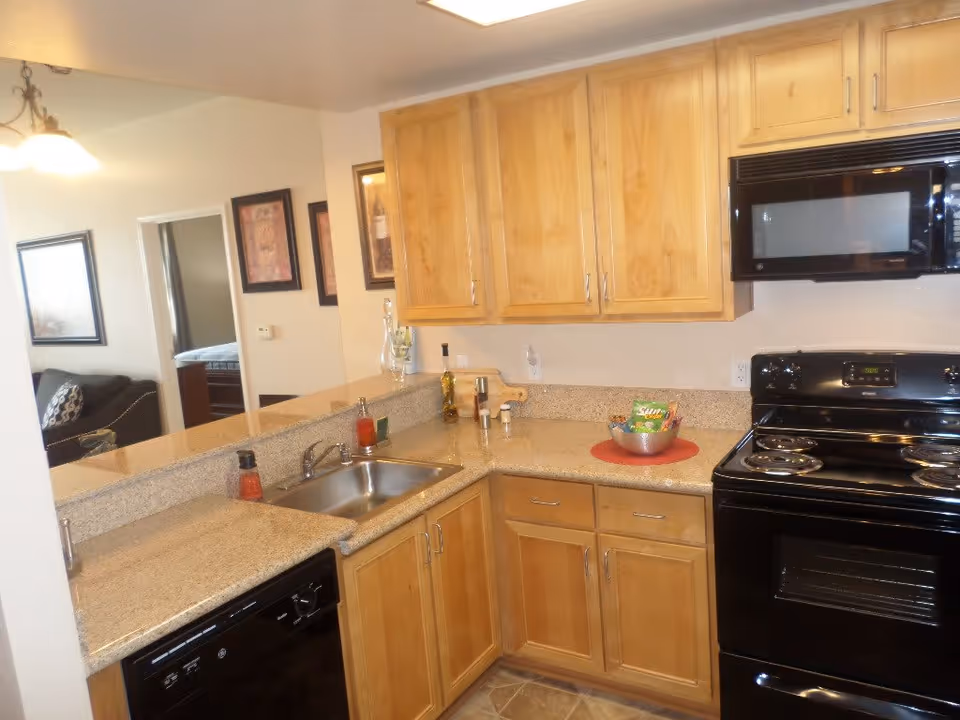 A kitchen area with wooden cabinets, a black electric stove and oven, a black microwave, a stainless steel sink, and a dishwasher. The countertop has a few items including a bowl with snacks and some bottles. In the background, a living room and a bedroom are partially visible through an open doorway.