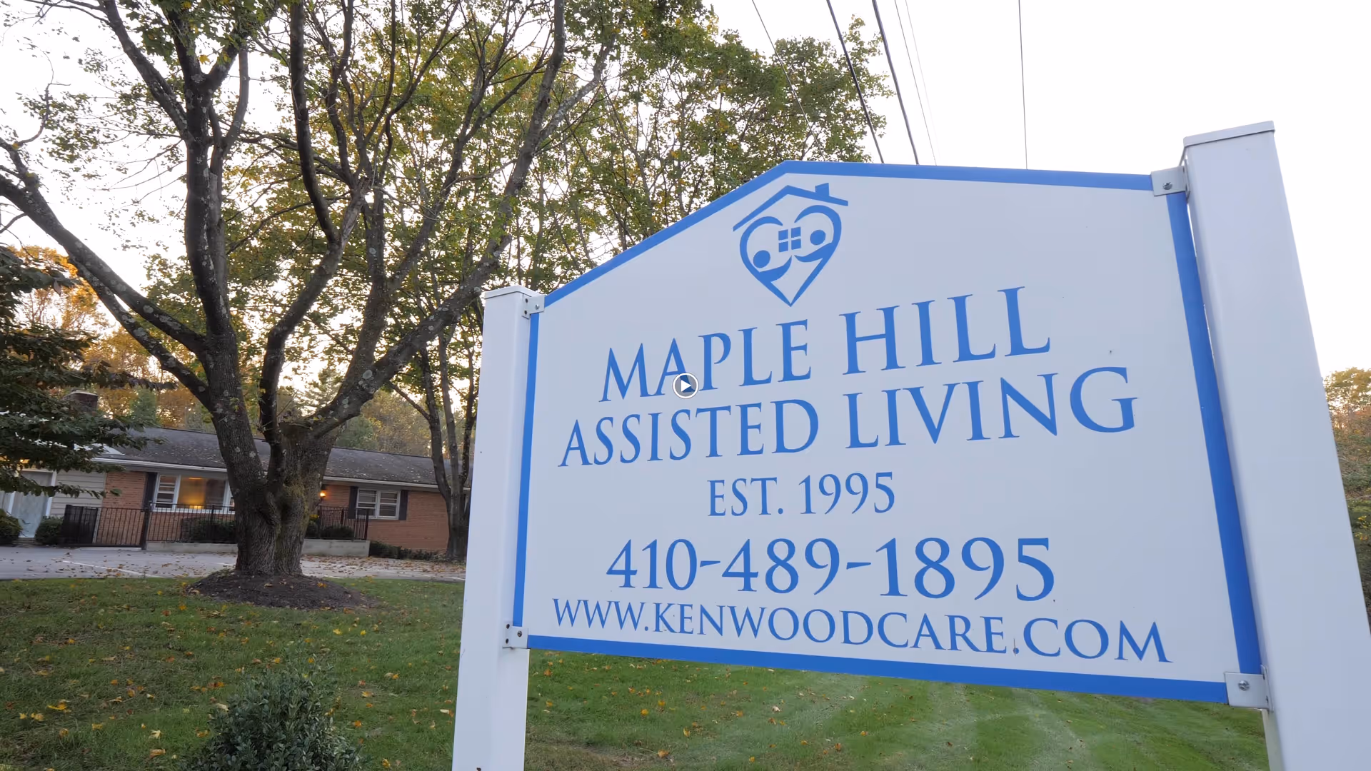A large white and blue sign for Maple Hill Assisted Living with contact information and website, set on a grassy area with trees and a single-story brick building in the background.