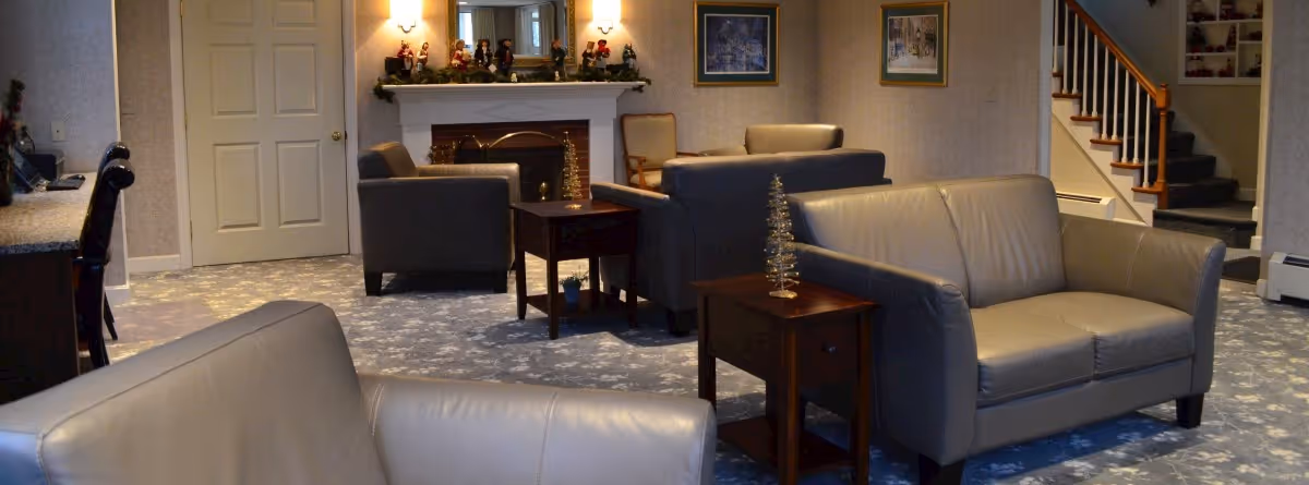 A cozy living room area in a nursing home with several gray leather sofas and armchairs arranged around wooden side tables. The room features a fireplace decorated with small figurines and garland, framed artwork on the walls, and a staircase leading upstairs. The carpet has a floral pattern, and the lighting is warm and inviting.