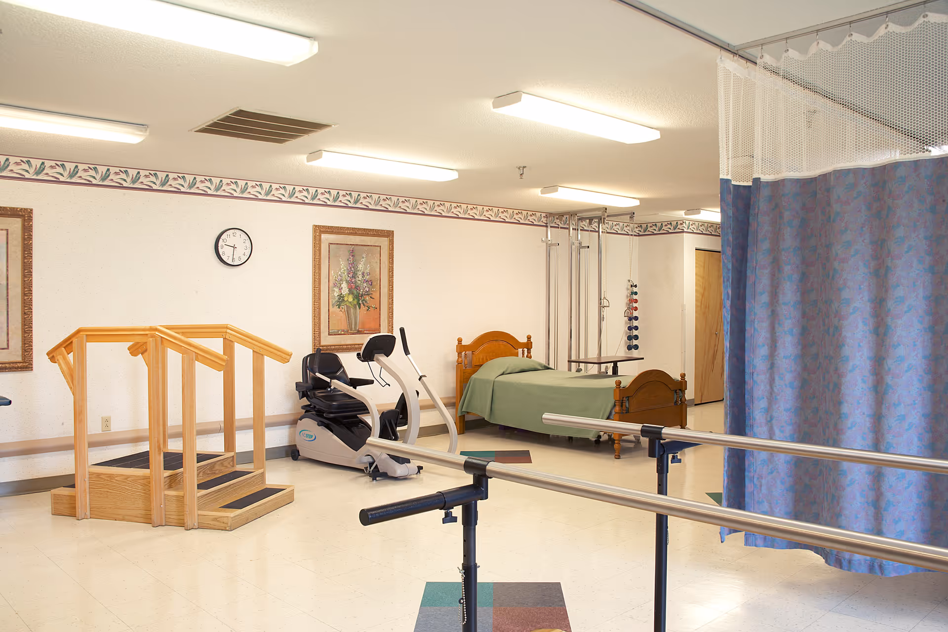 A rehabilitation or physical therapy room with parallel bars for walking exercises, a wooden step platform, a stationary exercise bike, and a hospital bed with a green blanket. The room has floral border wallpaper, framed floral artwork, and a clock on the wall. A blue privacy curtain is partially visible on the right side.