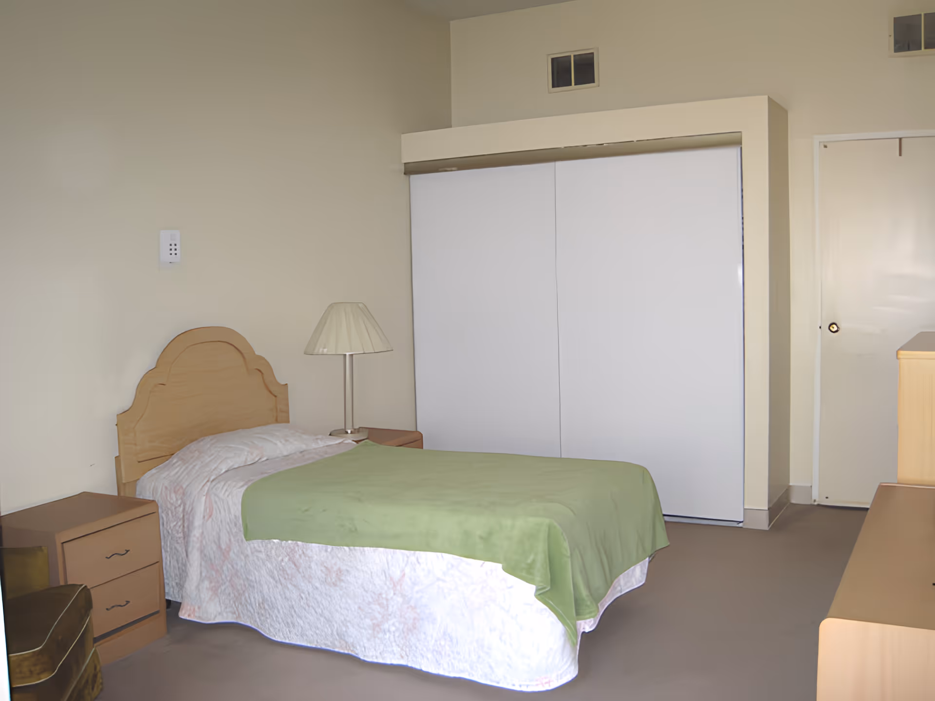A simple bedroom with a single bed covered with a white and pink bedspread and a green blanket. There is a wooden headboard, a nightstand with a lamp, a small dresser, a green armchair, and a large white closet with sliding doors. The walls are light-colored and there is a closed door in the background.