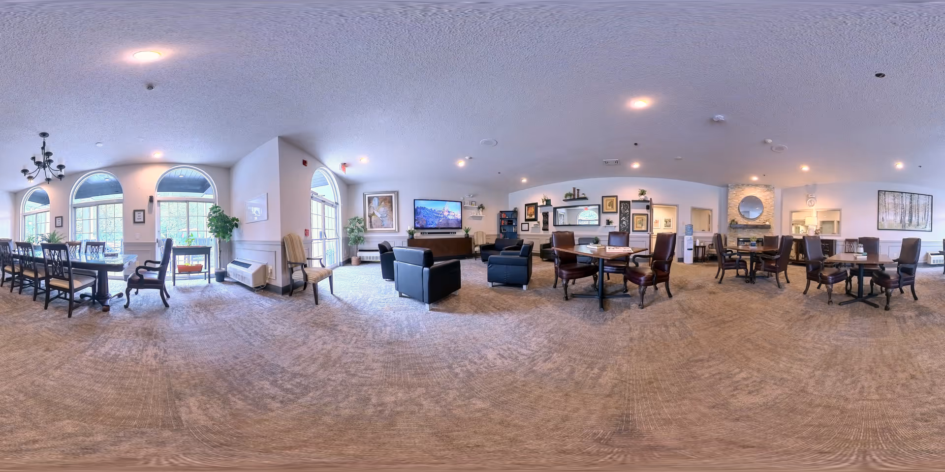A spacious senior living facility common area with multiple seating arrangements including dining tables with chairs, armchairs, and sofas. Large arched windows allow natural light to fill the room. The walls are decorated with framed artwork and plants. A flat-screen TV is mounted on one wall above a wooden cabinet. There is a fireplace with a round mirror above it, and the carpeted floor adds warmth to the space.