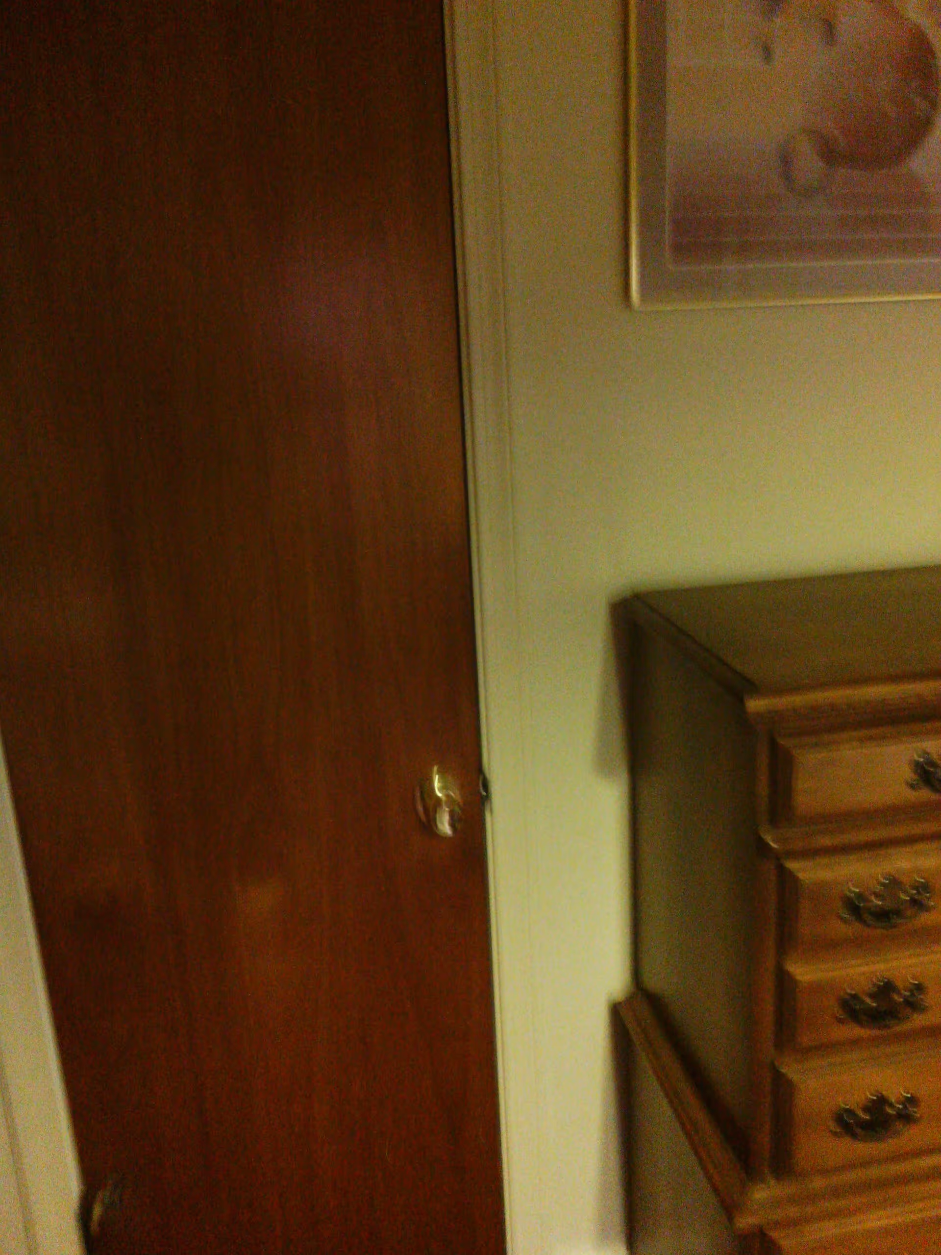 A wooden door with a brass-colored doorknob next to a wooden dresser with ornate handles. A framed picture is partially visible on the wall above the dresser.