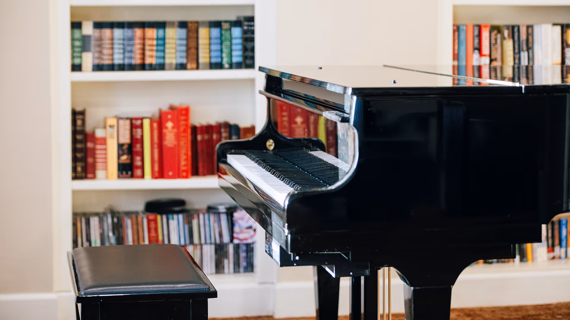 A black grand piano with a matching black piano bench in front of a white bookshelf filled with books and CDs in a cozy room.