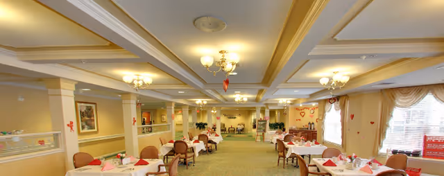 Bright dining room in a senior living facility with multiple set tables, chandeliers, columns, and large windows.