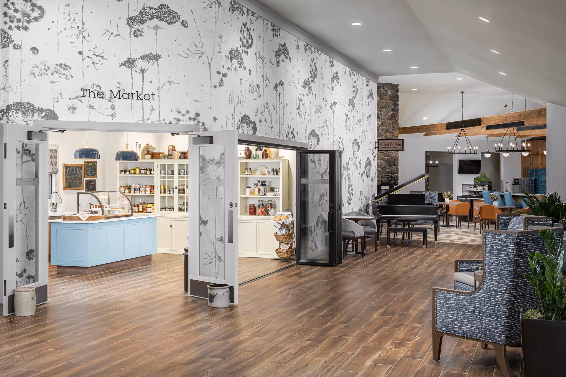 Interior view of a senior living facility showing a spacious common area with wooden flooring, patterned wallpaper, and a section labeled 'The Market' featuring shelves stocked with various items and a blue counter. The area also includes comfortable seating, a grand piano, and a lounge bar named 'Clock & Key' with modern lighting and decor.