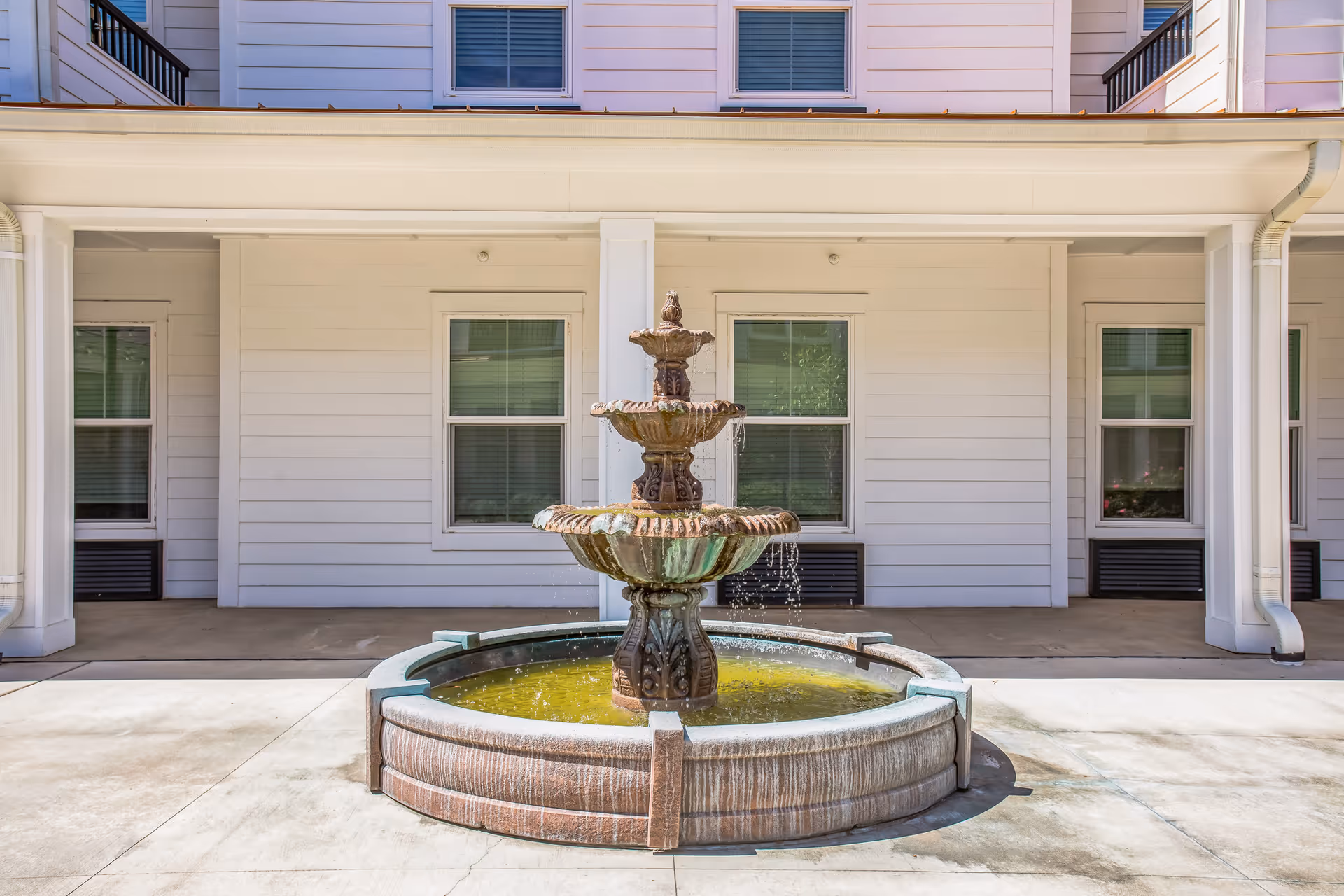 Outdoor courtyard area featuring a three-tiered stone water fountain with water flowing into a circular basin, set against the backdrop of a white building with multiple windows and a covered walkway.