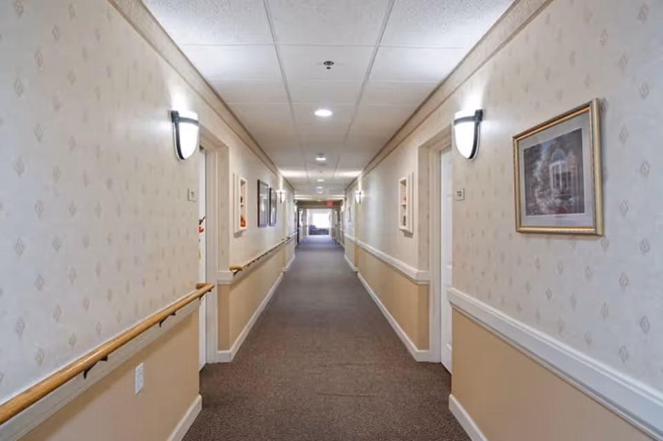 A long, well-lit hallway in a senior living facility with beige walls, carpeted floor, handrails on both sides, wall-mounted lights, framed pictures, and several doorways leading to rooms.