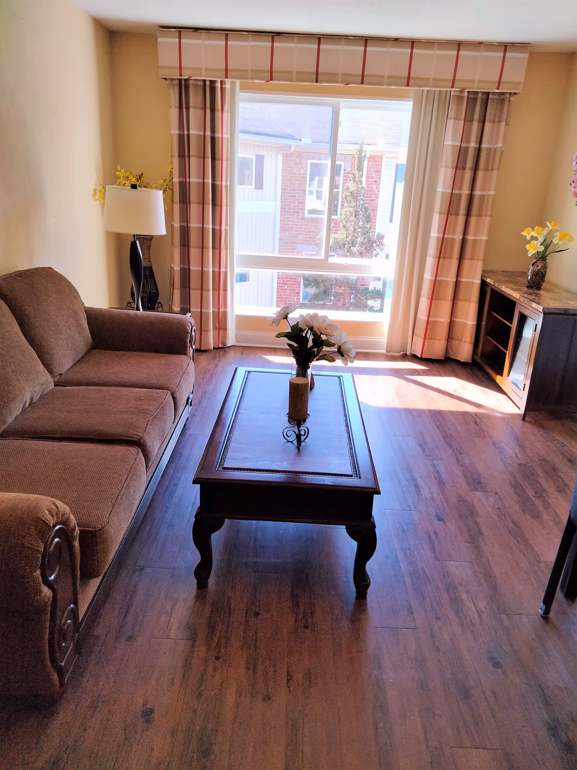 A cozy living room with a brown upholstered sofa on the left, a wooden coffee table with a vase of white flowers in the center, and a wooden cabinet with a vase of yellow flowers on the right. The room has wood flooring and a large window with plaid curtains letting in natural light.