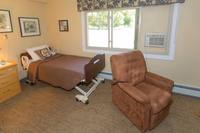 A small, neatly arranged room with a single hospital-style bed covered with a brown blanket and a decorative pillow. Next to the bed is a wooden nightstand with a lamp and some items on it. A large brown recliner chair is positioned near a window with a floral valance, and an air conditioning unit is installed in the window. The walls are light-colored and decorated with framed pictures.