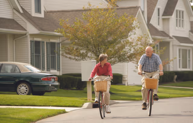 Two older adults ride bicycles with wicker baskets down a residential street in front of homes.