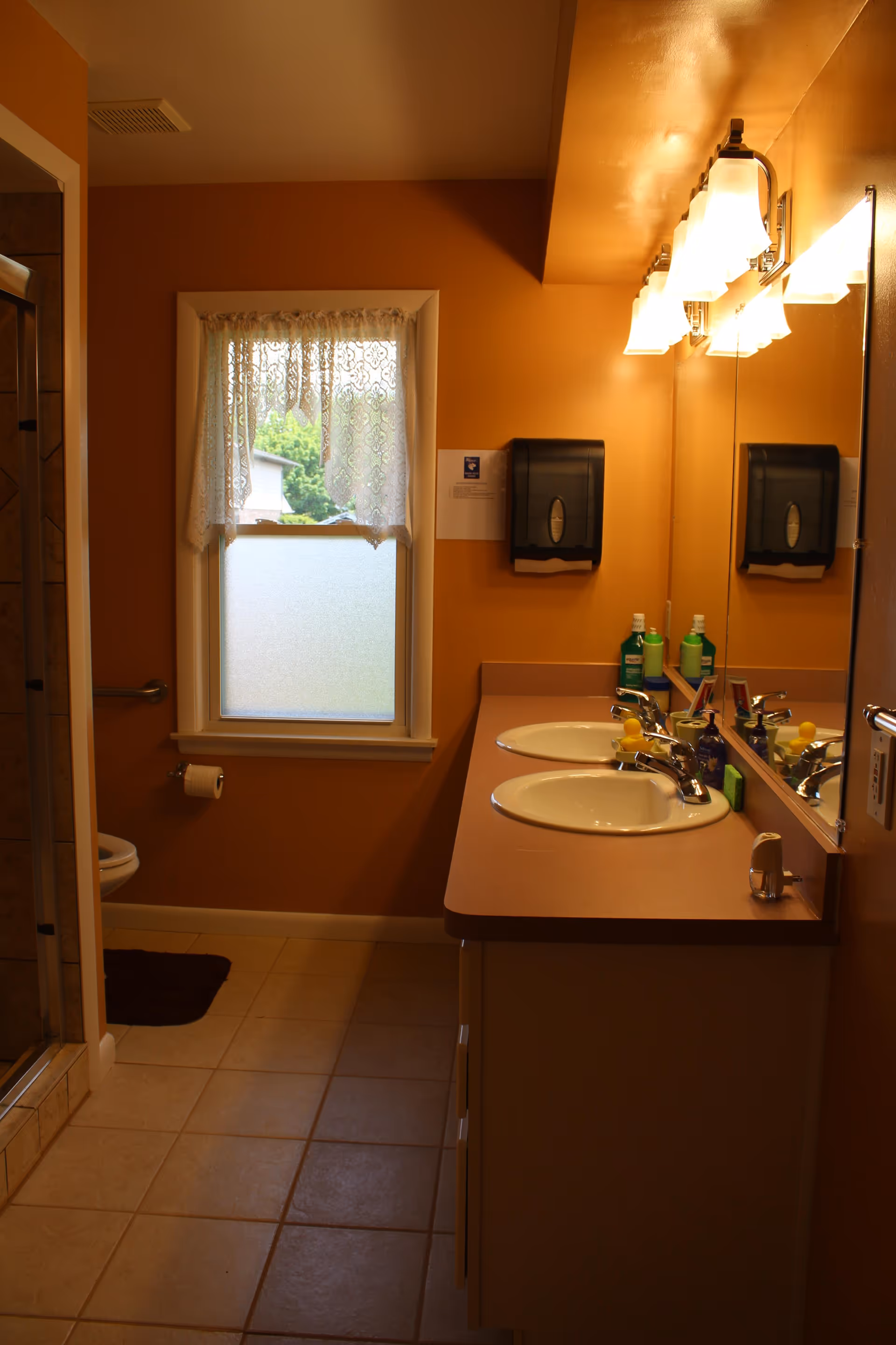 A bathroom with orange walls featuring a double sink vanity with a large mirror above it. There are two sinks with chrome faucets, various toiletries on the countertop, and a wall-mounted paper towel dispenser. A window with lace curtains lets in natural light. To the left, there is a toilet with a grab bar and a tiled shower area partially visible.