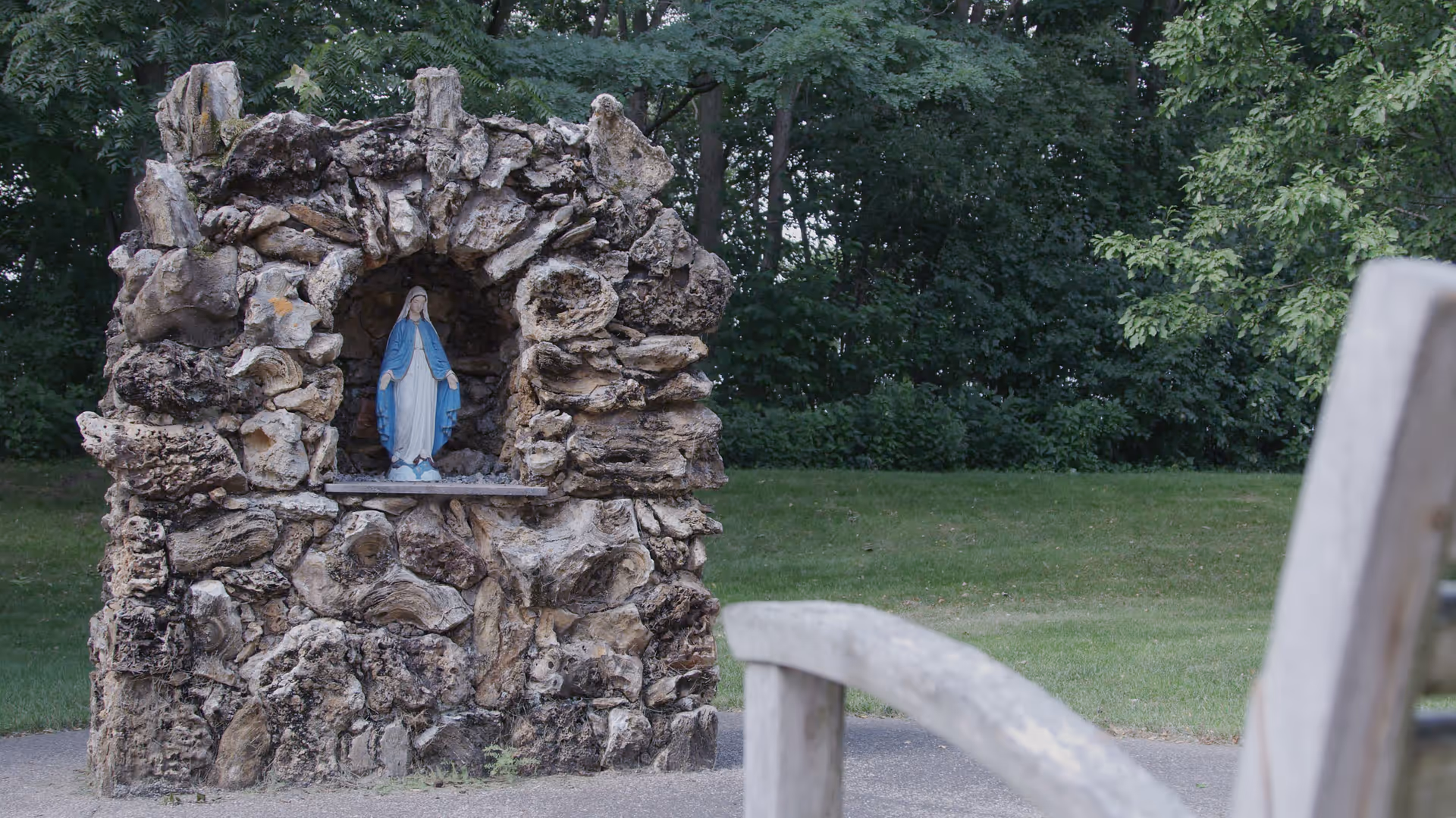 A small stone grotto holding a statue of the Virgin Mary with grass, trees and a wooden bench in the foreground.