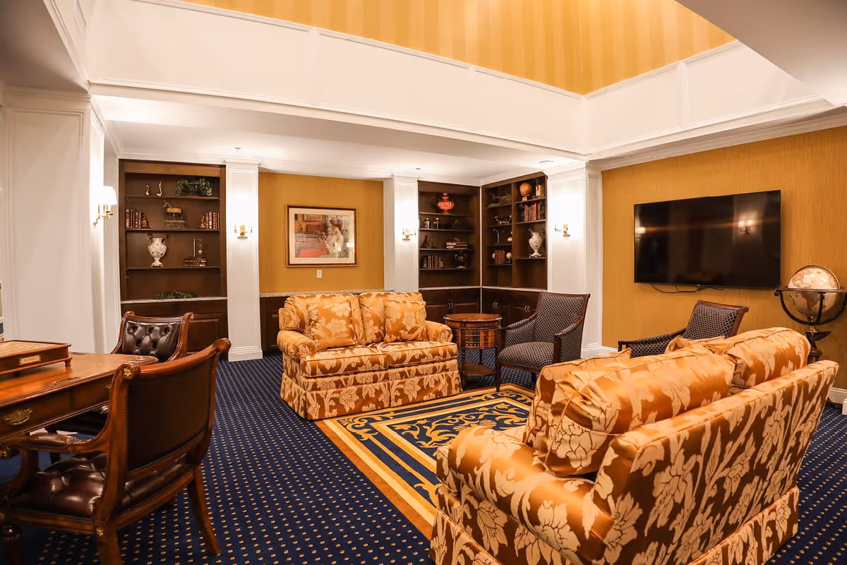 A cozy living room area with two patterned gold sofas, two dark upholstered armchairs, a wooden desk with leather chairs, built-in dark wood bookshelves, a wall-mounted flat screen TV, and a globe on a stand. The room has a blue carpet with a decorative gold and blue rug, warm yellow walls, and white trim.