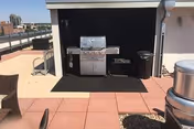 Rooftop patio area featuring a stainless steel gas grill under a small shelter, outdoor seating, and a city skyline in the distance.
