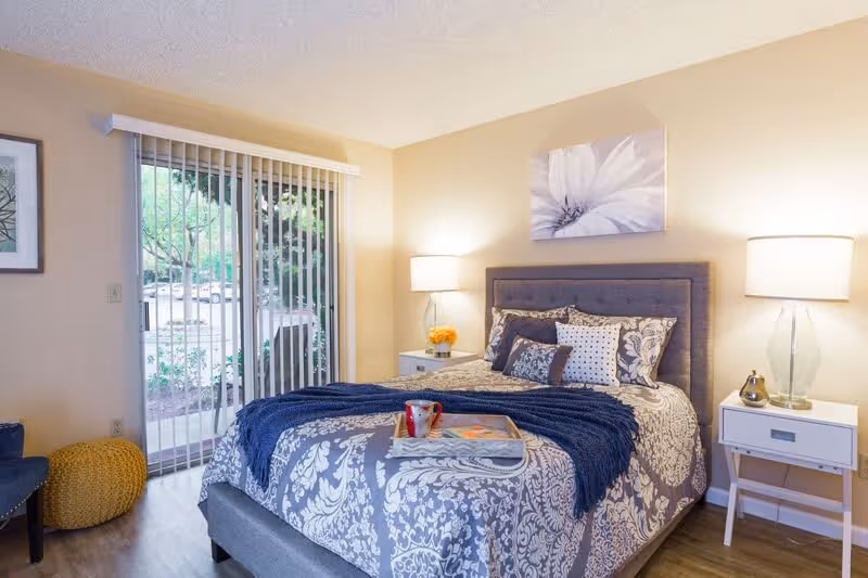 A cozy bedroom with a bed featuring a gray and white floral patterned comforter and multiple pillows. There is a blue throw blanket and a tray with a cup and book on the bed. Two white nightstands with lamps flank the bed. A large window with vertical blinds shows greenery outside. A yellow pouf and a blue chair are visible in the corner.