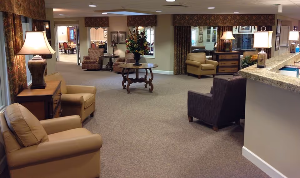 A spacious and well-lit common area in a senior living facility with several armchairs arranged around the room, a round wooden table with a floral arrangement in the center, table lamps on side tables, and a granite countertop on the right side. The room has carpeted floors, large windows with patterned curtains, and leads into a dining area in the background.