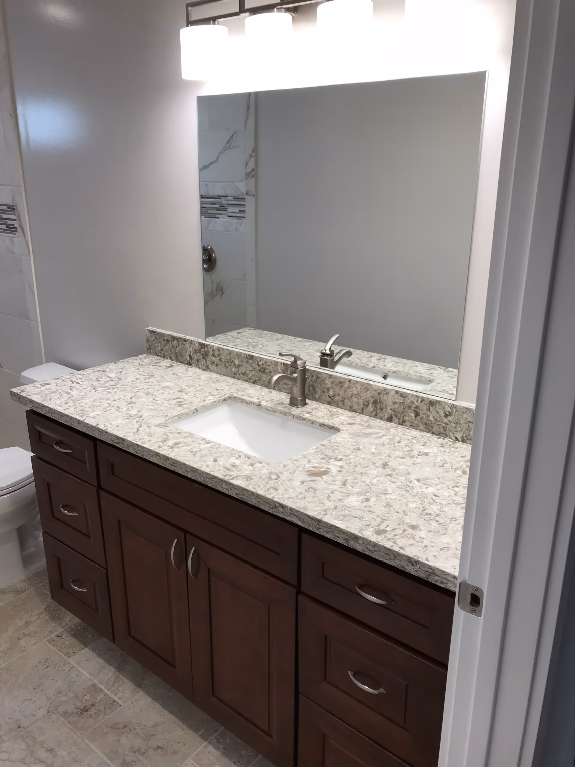 A bathroom vanity with a granite countertop, an under-mount sink, and a modern faucet. Above the vanity is a large rectangular mirror with three light fixtures mounted on the wall. To the left, part of a white toilet and tiled shower wall with decorative tile accents are visible.