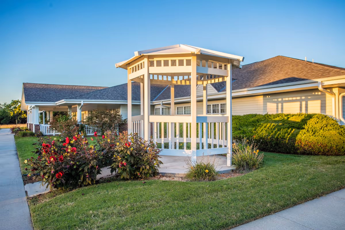 A white wooden gazebo surrounded by green grass, red flowers, and shrubs in front of a beige building with a brown shingled roof under a clear blue sky.