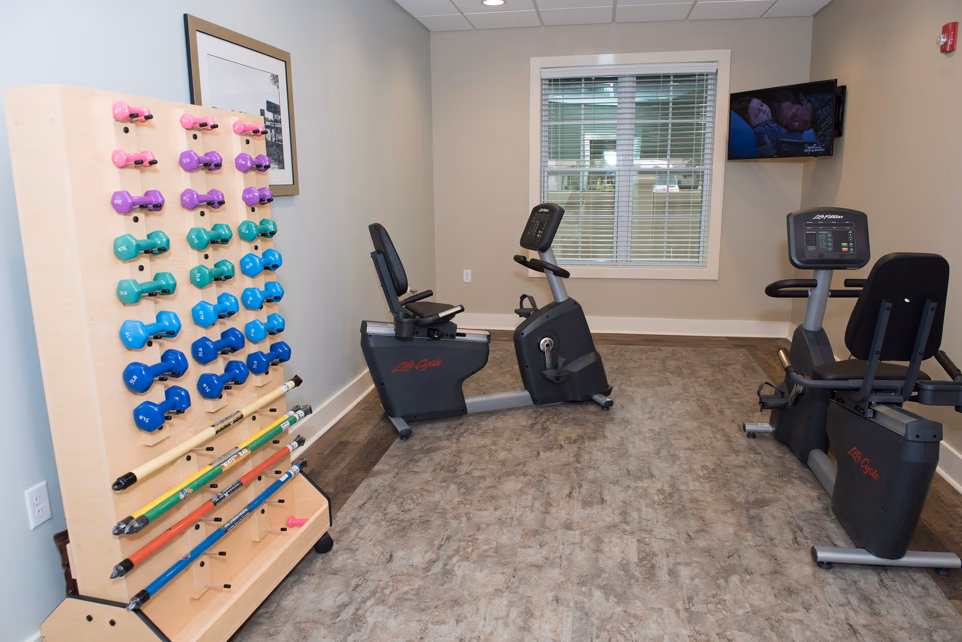 A small fitness room with two recumbent exercise bikes facing a wall-mounted TV. To the left, there is a wooden rack holding various colorful dumbbells and exercise bars. A window with blinds is on the back wall, and the floor has a textured, stone-like appearance.