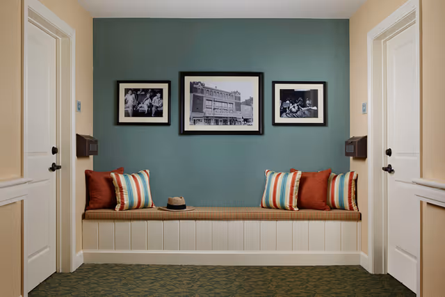 A cozy hallway seating area with a built-in bench featuring striped and solid red cushions and a hat placed on the bench. The wall behind the bench is painted teal and decorated with three black and white framed photographs. There are two white doors on either side of the bench, each with a black handle and a small mailbox attached to the wall beside them.
