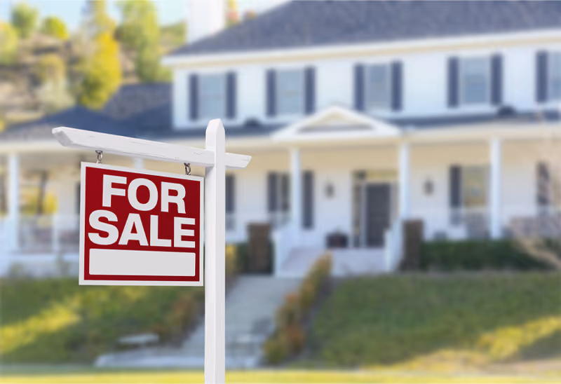 A red and white 'For Sale' sign in front of a large white house with a porch and multiple windows, surrounded by greenery.