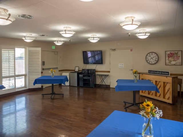 A spacious room with wooden flooring and several tables covered with blue tablecloths, each adorned with a small vase of flowers. The room features a wall-mounted TV, a piano with a sign on top, a clock, and framed artwork. Large windows with white shutters allow natural light to enter.