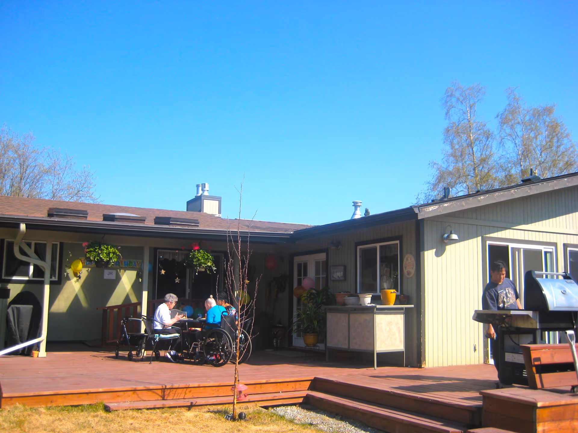 Outdoor wooden patio and deck beside a single-story assisted living building with several seated residents at a table and a person standing at a grill.