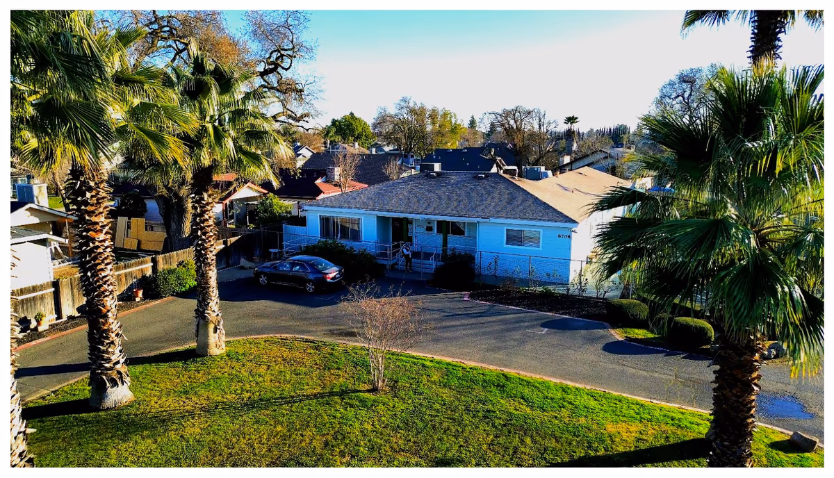 Exterior view of a single-story residential building with a dark roof and light blue walls, surrounded by palm trees and other greenery. A car is parked in the driveway, and a person is visible near the entrance. The scene is sunny with a clear sky.