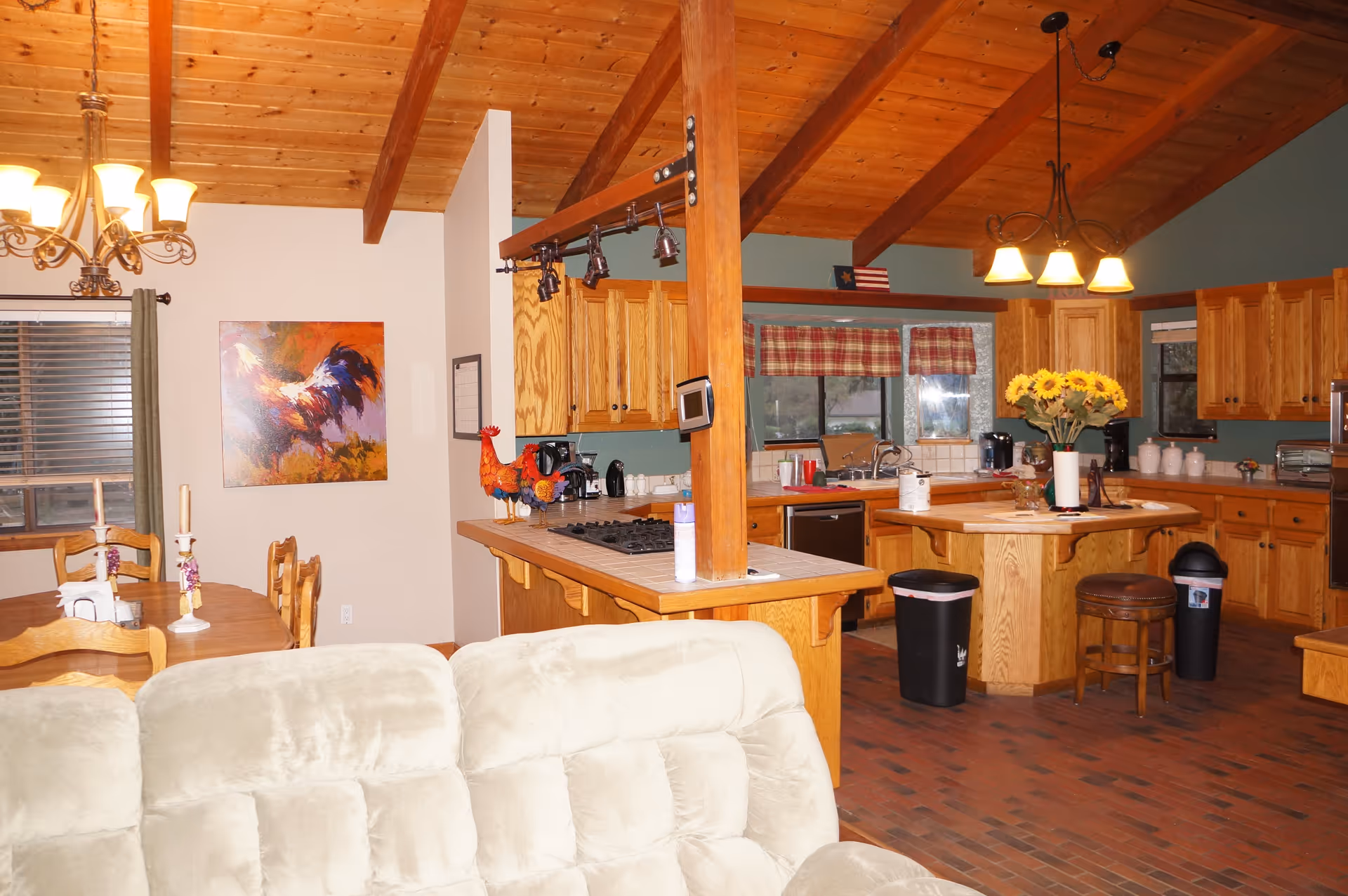 Interior view of a senior living facility showing a combined kitchen and dining area with wooden cabinets, a kitchen island with a vase of sunflowers, a dining table with chairs and candlesticks, a beige cushioned sofa in the foreground, and a wooden ceiling with exposed beams.