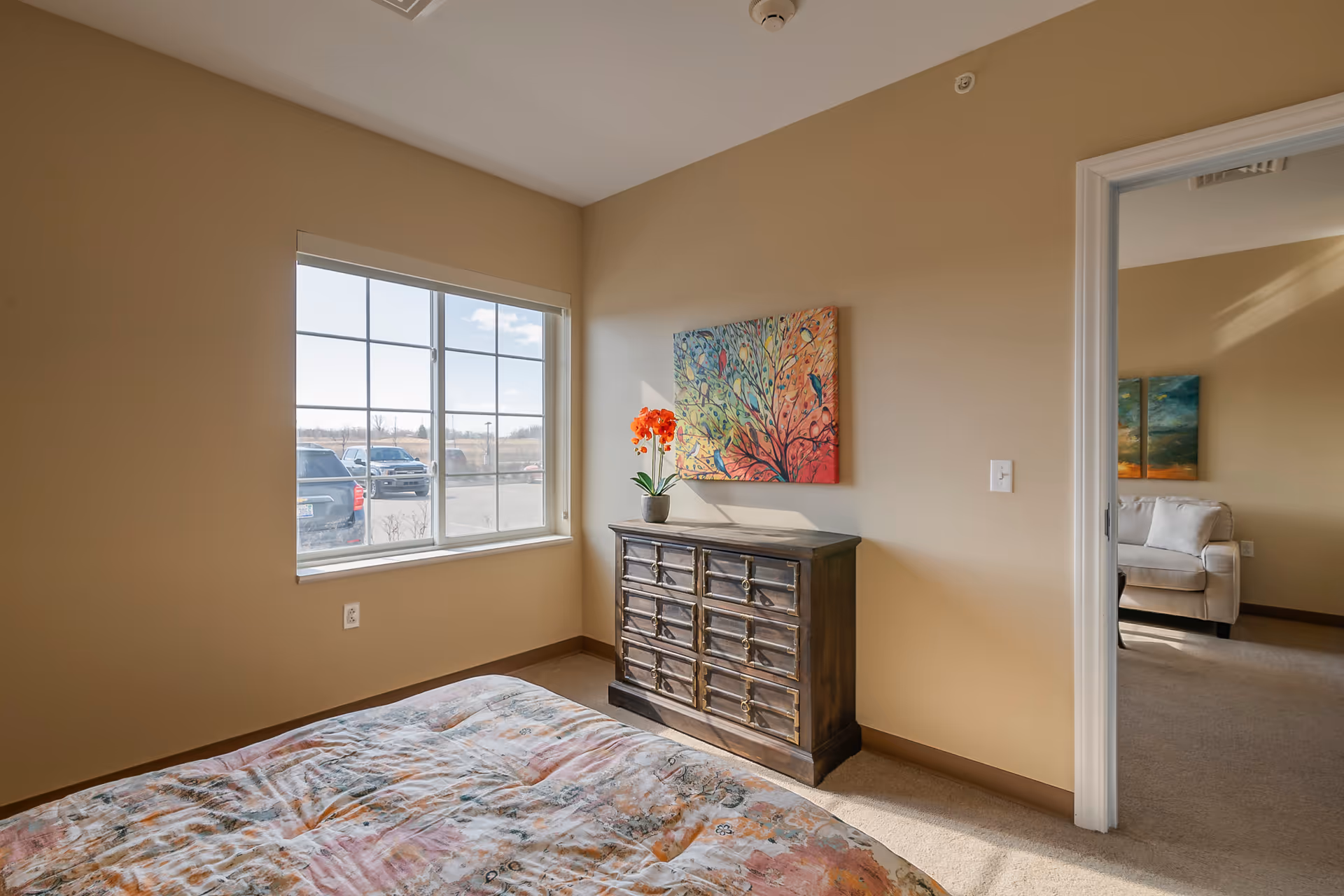 A bedroom with a window showing a parking lot outside. Inside the room, there is a bed with a colorful patterned bedspread, a dark wooden dresser with multiple drawers, and a vibrant painting of a tree with birds on the wall above the dresser. Through an open doorway, a beige sofa and additional artwork are visible in an adjacent room.