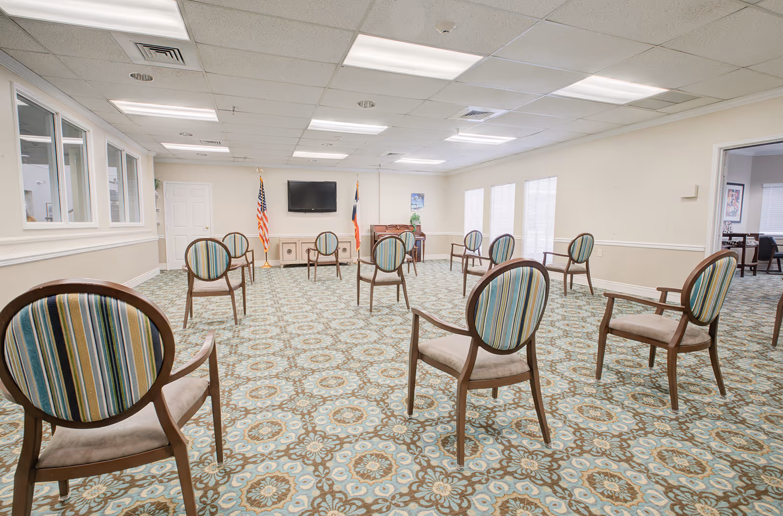 A spacious room with patterned carpet and several wooden chairs with striped upholstery arranged in a socially distanced manner. The room has a white ceiling with fluorescent lights, a wall-mounted TV, two flags, and a piano in the corner.
