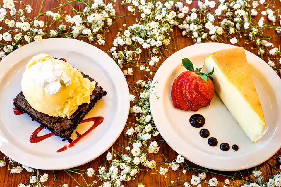 Two dessert plates on a wooden table decorated with small white flowers. One plate has a brownie topped with a scoop of vanilla ice cream and whipped cream, drizzled with red sauce. The other plate has a slice of cheesecake with a sliced strawberry and chocolate sauce dots.