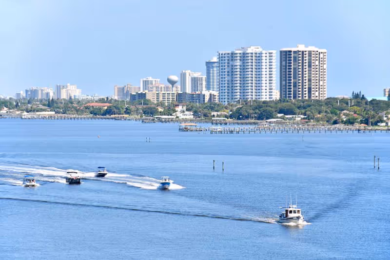 View of a waterfront city skyline with several high-rise buildings in the background and multiple boats speeding across the water in the foreground under a clear blue sky.