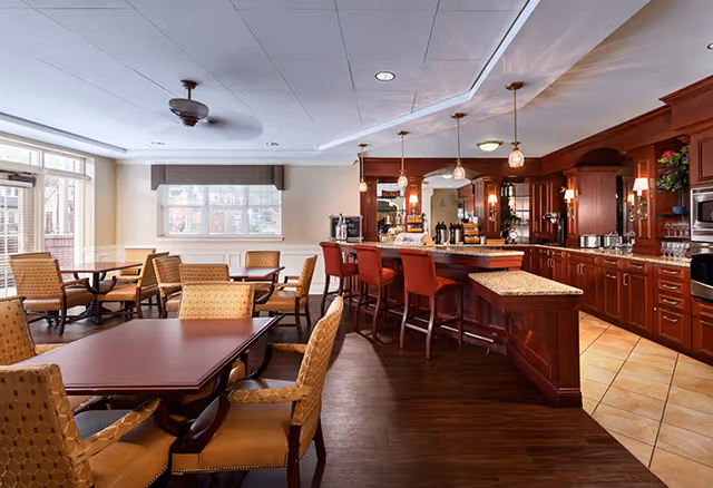 A spacious dining area with multiple tables and cushioned chairs, adjacent to a kitchen area featuring wooden cabinetry, a countertop bar with red bar stools, pendant lighting, and kitchen appliances. Large windows allow natural light to fill the room.