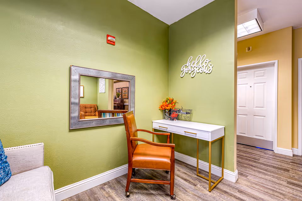 An interior corner of a senior living facility with green walls and wood flooring. There is a white console table with gold legs against the wall, decorated with a small bouquet of orange flowers and a wire basket. Above the table, a decorative sign reads 'hello gorgeous'. A brown leather chair with wooden arms is placed in front of the table. A large rectangular mirror with a metallic frame hangs on the adjacent wall, reflecting part of the room including a brown couch and framed artwork. A beige couch with a blue cushion is partially visible on the left side. In the background, a hallway with beige walls and a white door is visible.