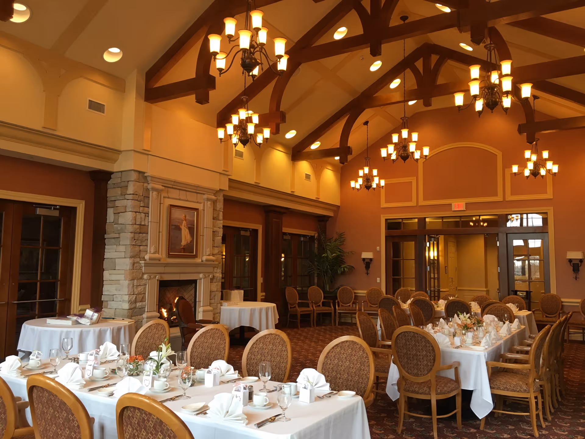 A spacious dining room with high vaulted ceilings featuring wooden beams and multiple chandeliers. The room has several tables covered with white tablecloths, set with plates, glasses, napkins, and silverware. There is a stone fireplace with a framed painting above it on one wall, and large windows and doors along the side. The room has warm lighting and a cozy, elegant atmosphere.