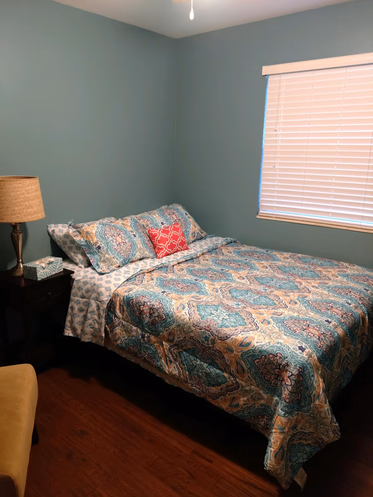 A bedroom with a bed covered in a patterned blue, teal, and beige comforter with matching pillows and a small red decorative pillow. There is a wooden nightstand with a lamp and a box of tissues next to the bed. The walls are painted light blue, and there is a window with closed white blinds. The floor is wooden.