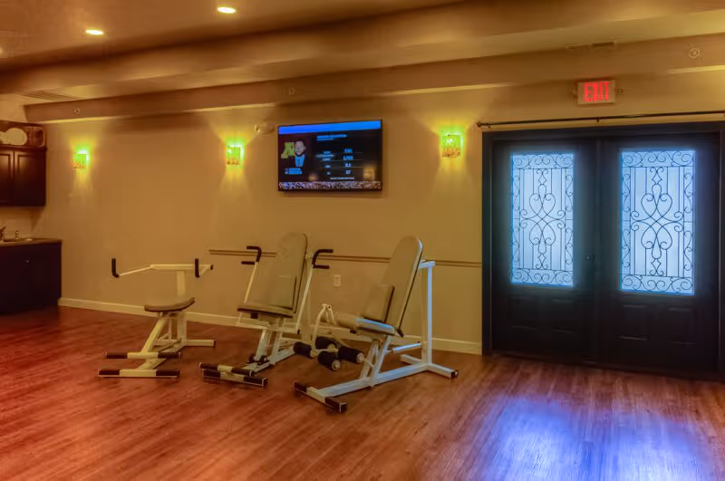 An interior room with wooden flooring featuring two exercise machines positioned against a beige wall. Above the machines is a mounted flat-screen TV displaying a program. The room is softly lit by wall sconces, and there is a set of double doors with decorative glass panels on the right side under an illuminated exit sign.