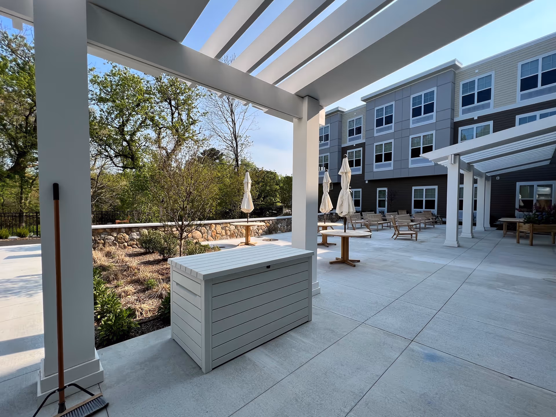 Outdoor patio area at The Residence at Natick South featuring a covered seating area with white pergola beams, several round tables with closed umbrellas, wooden chairs, and a stone wall with greenery and trees in the background under a clear sky.