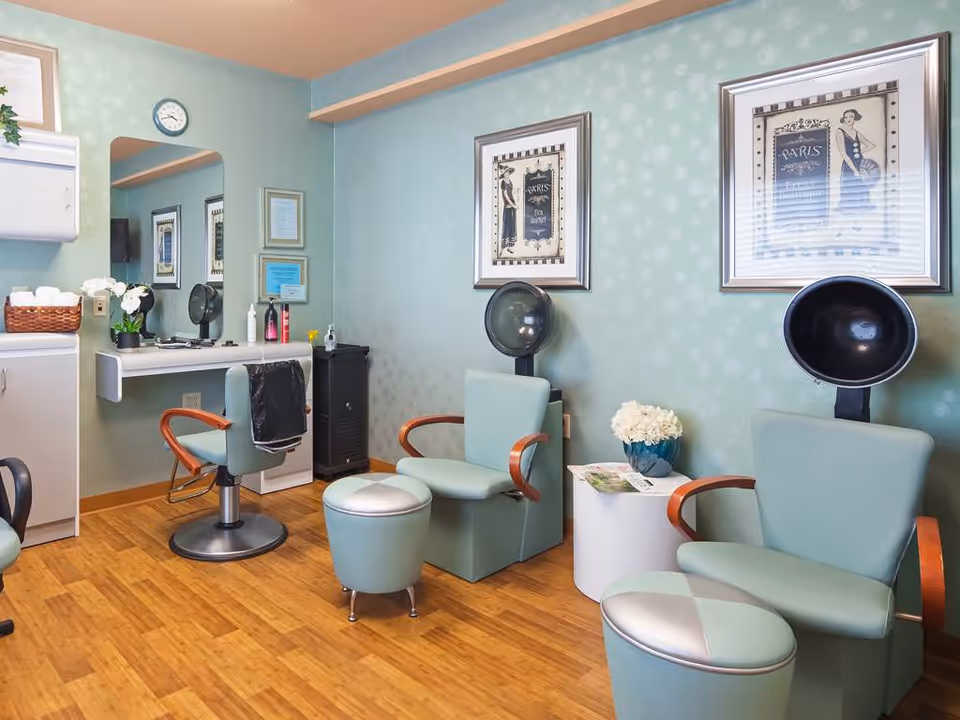 Interior of a salon area in a senior living facility with light green walls and wooden flooring. The room features two vintage hair dryers, two light green salon chairs with wooden armrests, a small round white table with a flower vase and magazine, a wall-mounted mirror, and a salon workstation with a chair and hair care products. Two framed Paris-themed artworks hang on the wall.