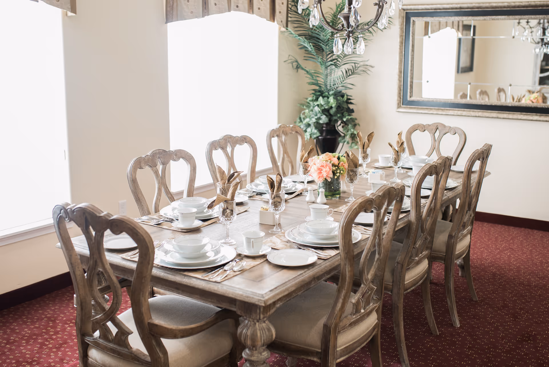 A dining room with a wooden table set for eight people. The table is arranged with white plates, bowls, cups, silverware, and glasses with folded napkins. A floral centerpiece and salt and pepper shakers are placed in the middle. The room has a red carpet, two large windows with curtains, a large mirror on the wall, and a potted plant in the corner.