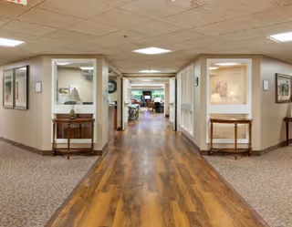 Interior hallway of a senior living facility with wood flooring in the center and carpeted sides, beige walls, and ceiling tiles. There are small tables with lamps and framed pictures on the walls. The hallway leads to a common area with seating and large windows in the background.