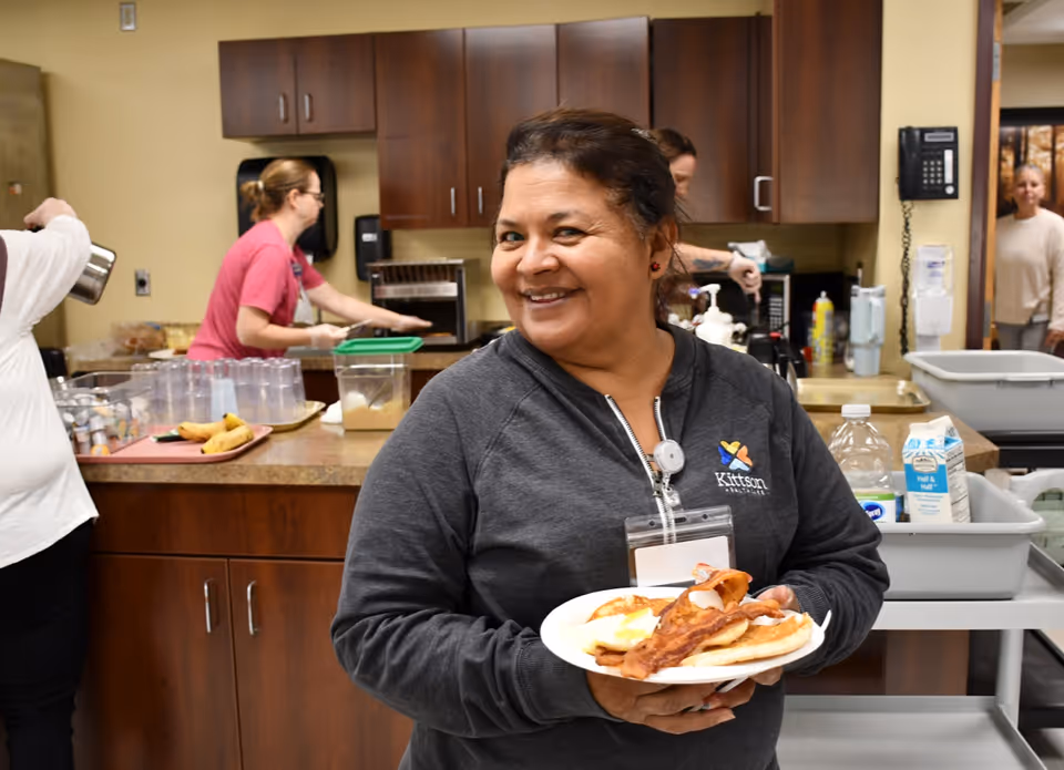 A smiling staff member holds a plate of bacon, eggs and pancakes in a facility kitchen with other workers and food prep in the background.